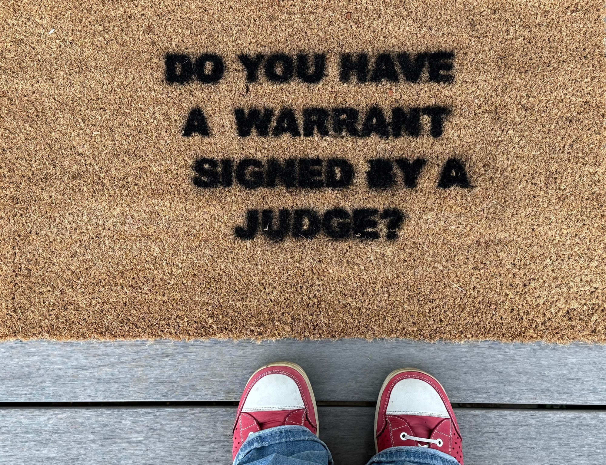 A brown door mat spray painted with black lettering reads "Do you have a warrant signed by a judge?"