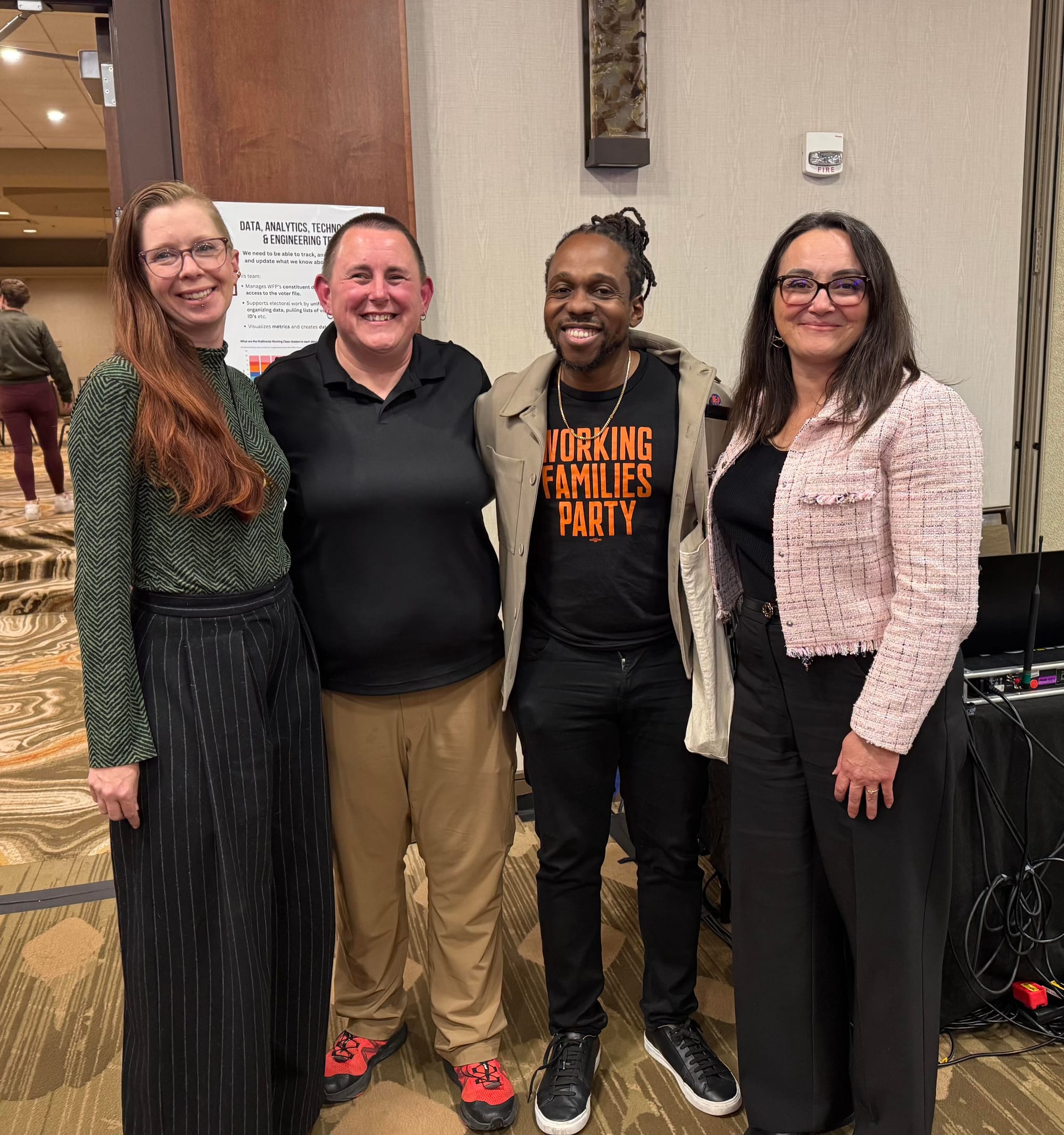 Four people stand in a hotel conference area. They are smiling and facing the camera.