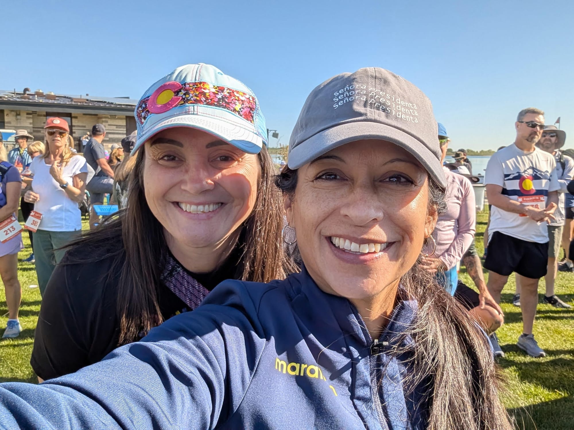 Nicole and Marta are standing on the grass in a crowd. The Boulder Reservoir is behind them.