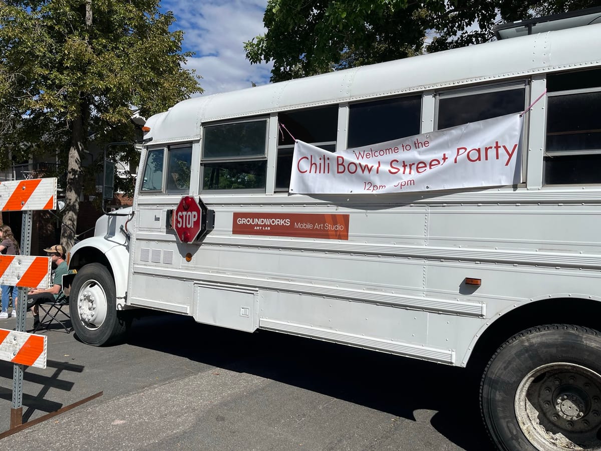A white bus blocks a street. An orange sign on the bus reads "Groundworks Art Lab Mobile Art Studio."