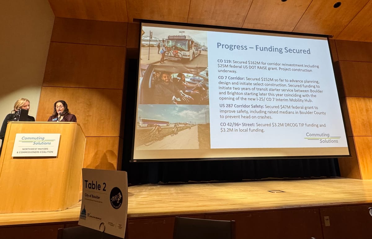 Two women stand at a podium. A presentation slide highlights 2025 transportation funding wins.