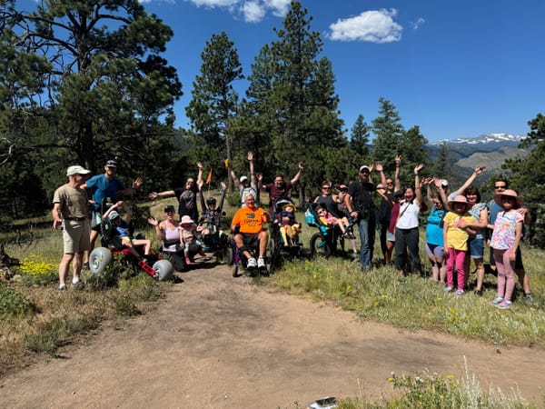 A group of people of all abilities celebrates their togetherness on a trail overlooking snow-covered mountains.