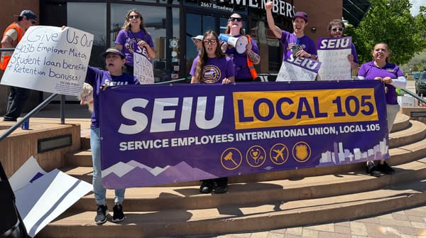 Seven people wearing purple t-shirts stand on a street corner holding their SEUI Local 105 union banner.