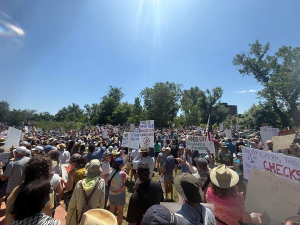 A sea of people stands under a blue sky in Boulder's civic area. Some hold protest signs.