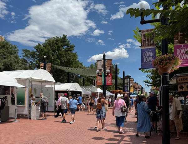 Visitors walk past art tents on Pearl Street during a sunny festival, with flower baskets and ‘Love the Local Downtown’ banners overhead.