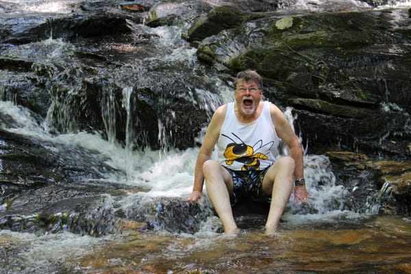 An elderly man sits in a rushing creek, laughing with surprise as cold water splashes around him.
