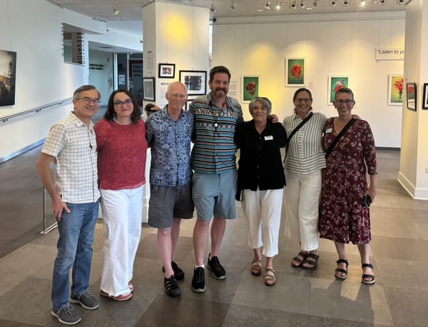 Seven people pose in a modern art gallery with framed artwork on the wall behind them.