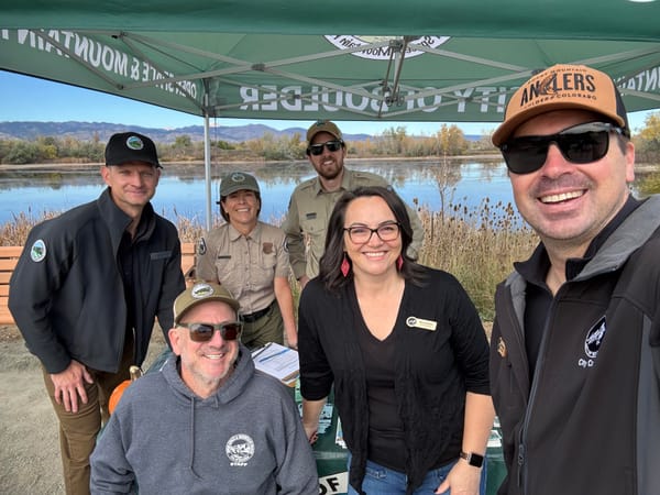 Six people smile under a "City of Boulder" tent with a pond and the Front Range in the background.