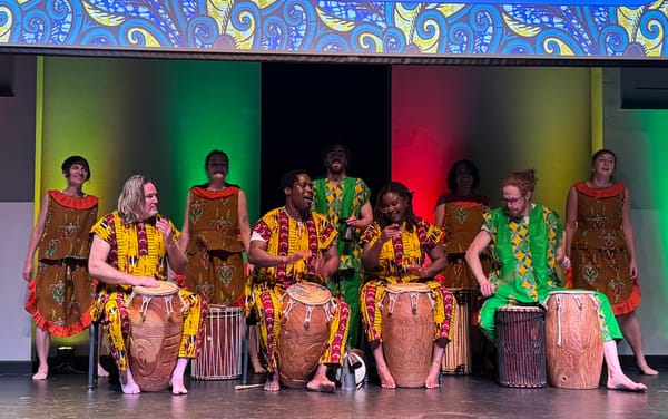 Musicians and dancers in vibrant African attire perform with hand drums under colorful stage lighting.